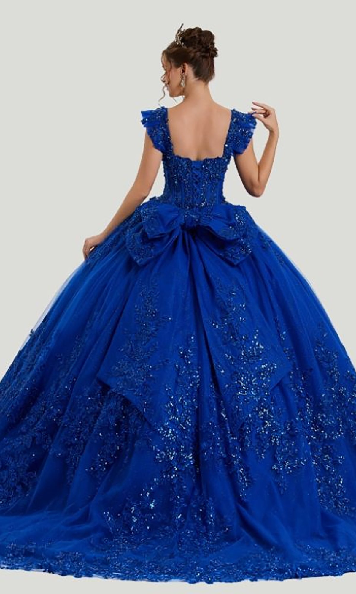 A woman wears the Quinceanera Dress 26410 By Stella Couture, an ornate royal-blue ball gown featuring a dramatic bow, sequined floral accents, and a stunning backless design. The voluminous skirt and exquisitely embellished bodice make this dress perfect for making a statement at any quinceañera celebration.
